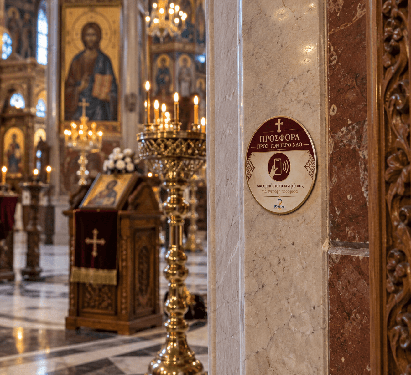 Targa per offerte contactless su una colonna di marmo all'interno di una chiesa, con icone e candele accese sullo sfondo